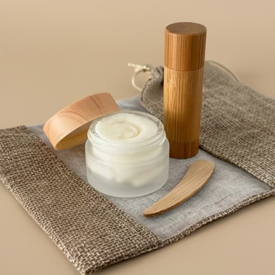 Skincare products including a jar, wooden applicator, and bamboo container on a textured surface.